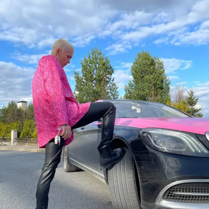 Даня Милохин в розовой куртке с ногами на колесах своего автомобиля