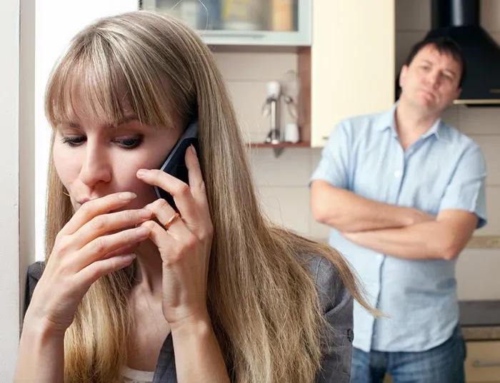 Жена разговаривает по телефону, муж скрестил руки и смотрит на неё с недовольным видом