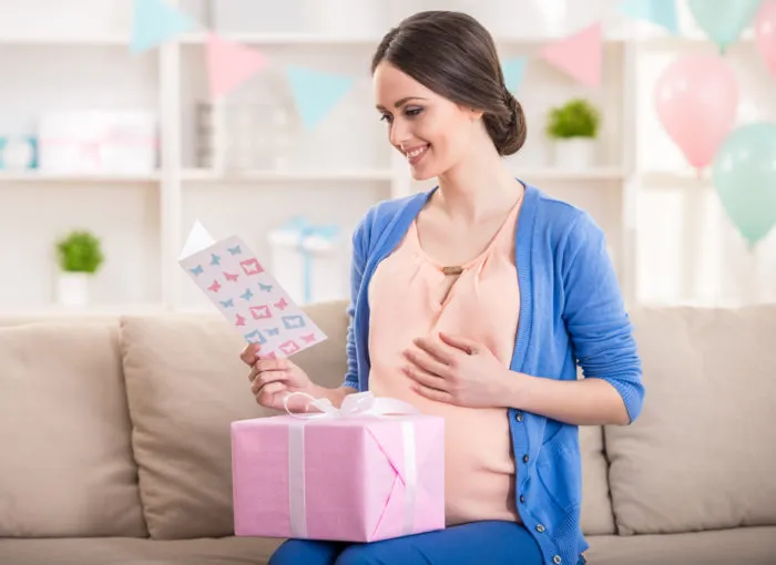 Smiling young pregnant woman is looking at the greeting card and holding a gift box while sitting on the couch.